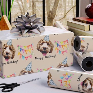 May include: A rectangular gift box wrapped in beige paper featuring a repeating pattern of dogs wearing blue polka-dot party hats, colorful bunting flags, and the text Happy Birthday, topped with a large silver metallic gift bow.