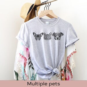 May include: A gray t-shirt with a black and white illustration of a dog, cat, and dog. The t-shirt is on a hanger and has a white background.