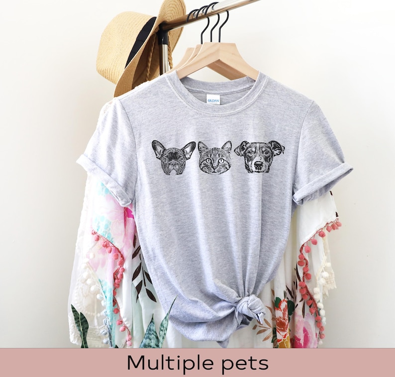 May include: A gray t-shirt with a black and white illustration of a dog, cat, and dog. The t-shirt is on a hanger and has a white background.