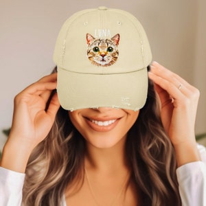 May include: A beige distressed baseball cap featuring a graphic illustration of a brown tabby cat face with green eyes and the name LUNA printed in white capital letters above the cat's head. The cap has a worn, frayed edge on the brim.