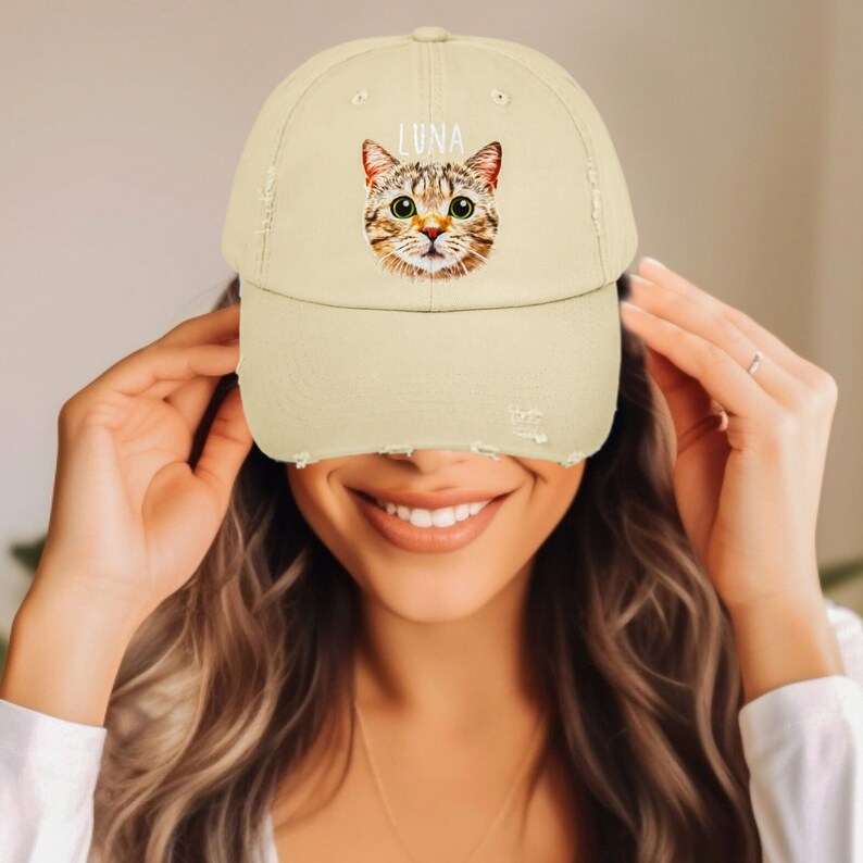 May include: A beige distressed baseball cap featuring a graphic illustration of a brown tabby cat face with green eyes and the name LUNA printed in white capital letters above the cat's head. The cap has a worn, frayed edge on the brim.