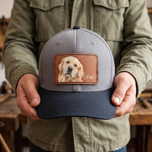 May include: A grey baseball cap with a dark navy blue brim and a rectangular brown leather patch on the front. The patch features a detailed illustration of a golden retriever dog and the name Charlie written in white cursive script.