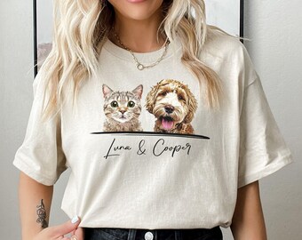 May include: A cream-coloured short-sleeve t-shirt featuring a graphic print of a brown tabby cat and a golden-brown doodle dog. Below the animals, a black horizontal line sits above the cursive black text that reads Luna & Cooper.