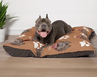 May include: A rectangular pet bed with a brown top surface featuring a repeating pattern of white paw prints and a dog's face with the name MAX printed in white. The sides of the bed are dark brown, and a gray dog is lying on the center of the bed.