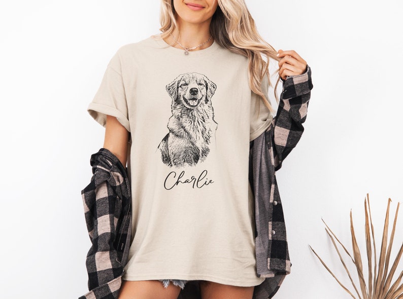 Beige t-shirt featuring a custom sketch-style portrait of a dog with the name 'Charlie' in cursive. Styled with a plaid shirt for a casual look, this personalized shirt makes a thoughtful gift for pet lovers or dog parents.