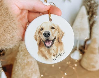 May include: A round white ceramic ornament featuring a detailed illustration of a golden retriever dog with its mouth open. The name Charlie is written in black cursive script below the dog. A gold string is attached to the top for hanging.