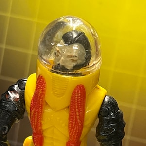 May include: Yellow and black action figure with a clear plastic helmet. The figure has red details on the chest and black arms.