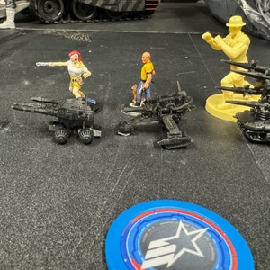 May include: A blue die with a white star and the number 2 on each side.  Three black plastic miniature vehicles with a yellow plastic miniature figure standing next to one of the vehicles.  A yellow plastic miniature figure with a hat standing next to a black plastic miniature vehicle with multiple missiles.  A blue and red circular token with a white star in the center.