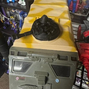 Op de afbeelding: Een speelgoedvoertuig met militair thema met een camouflagepatroon op het bovenste gedeelte en een donkergroen onderste gedeelte. Een zwart, cirkelvormig onderdeel met een handvat bevindt zich bovenop. Het voertuig heeft een gedetailleerd ontwerp met zichtbare panelen en markeringen.