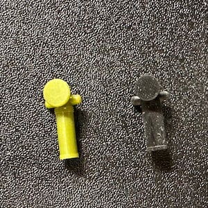 May include: Two small plastic figurines, one yellow and one black. The yellow figurine is shaped like a person holding a cylindrical object. The black figurine is shaped like a person with a round head and a cylindrical body.
