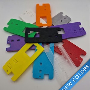 Puede incluir: Una colección de 10 fundas de teléfono de plástico en varios colores, incluyendo amarillo, azul, rojo, morado, verde, naranja, blanco, gris y negro. Las fundas tienen una forma rectangular con un recorte para la lente de la cámara del teléfono. El texto "NEW COLORS" es visible en la esquina inferior derecha de la imagen.