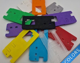 May include: A collection of 10 plastic phone cases in various colors, including yellow, blue, red, purple, green, orange, white, gray, and black. The cases have a rectangular shape with a cutout for the phone's camera lens. The text "NEW COLORS" is visible in the bottom right corner of the image.