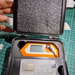 Könnte beinhalten: Geöffnete schwarze Box mit Schaumstoffauskleidung, die ein orange-weißes elektronisches Gerät mit Bildschirm und einen kleinen Metallschlüsselanhänger enthält. Eine Leiterplatte ist ebenfalls sichtbar. Die Box liegt auf einer grünen Schneidematte.