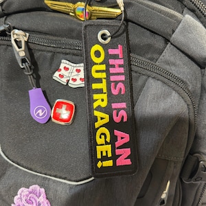 May include: A rectangular black fabric keychain tag featuring the embroidered text THIS IS AN OUTRAGE! in bright pink and yellow thread. The tag includes a silver metal grommet at the top and is attached to a black cord loop with a metal fastener.