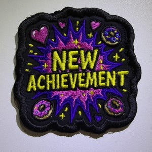 May include: A black embroidered patch featuring the yellow text NEW ACHIEVEMENT centered over a purple and pink starburst design. The patch is decorated with small pink hearts, yellow stars, and two small donuts with pink frosting and sprinkles.