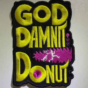 May include: A black embroidered patch featuring the yellow text GOD DAMNIT DONUT. The design includes a small pink graphic of a person running, three small pink skulls, and a pink center in the letter O, all outlined in black thread.