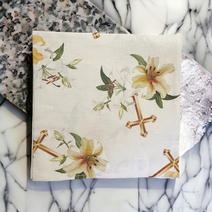 May include: Square, off-white handkerchief with a floral and cross design. The handkerchief features yellow and white lilies with green leaves and stems. Gold-colored crosses are also printed on the fabric. The handkerchief is folded and placed on a marble surface.