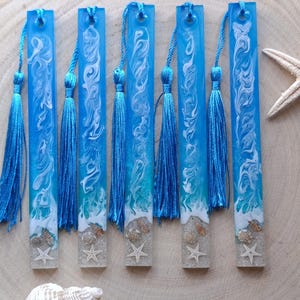 May include: Six resin bookmarks with a beach theme. Each bookmark is a vibrant blue with white swirls, resembling ocean waves. They feature blue tassels and include seashells and starfish embedded in a sandy base. The bookmarks are approximately 6 inches long.