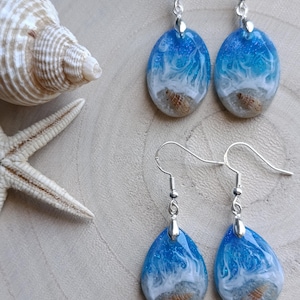 May include: Two pairs of drop earrings featuring resin pendants with a beach theme. The top pair is oval-shaped, and the bottom pair is teardrop-shaped. Both feature blue and white swirling waves, glitter, and small embedded seashells.
