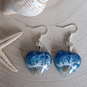 May include: A pair of heart-shaped drop earrings featuring a resin design that mimics ocean waves crashing onto a sandy shore. Each earring contains a small, embedded seashell and is suspended from a silver-toned metal hook for pierced ears.