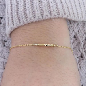Peut inclure: Un bracelet en chaîne dorée avec de petites perles dorées et argentées. Le bracelet est porté au poignet.