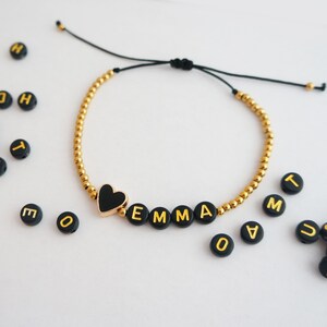 May include: A gold beaded bracelet with a black heart charm and black beads spelling out the name "EMMA".