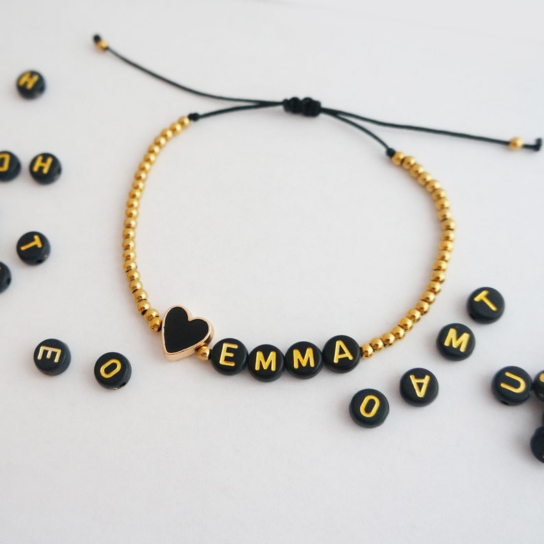 May include: A gold beaded bracelet with a black heart charm and black beads spelling out the name "EMMA".