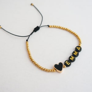 May include: A black and gold beaded bracelet with a black heart charm and the name "EMMA" spelled out in gold letters.