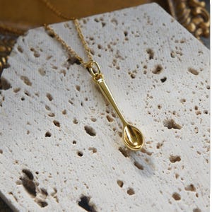 May include: A gold-colored spoon pendant necklace. The spoon-shaped charm is suspended from a delicate chain. The necklace is displayed on a textured, off-white surface, creating a contrast with the shiny gold. The spoon charm is approximately 1 inch in length.