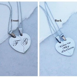 May include: Two silver necklaces with heart-shaped pendants. One pendant has the letters "F" and "G" engraved on it, while the other pendant has the text "You're My Person" engraved on it.