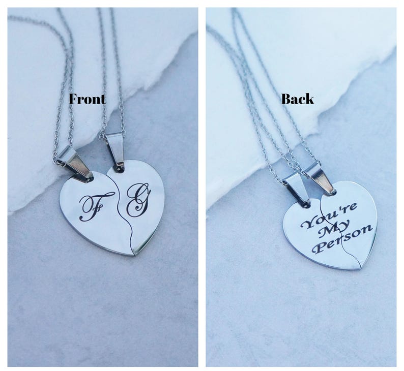 May include: Two silver necklaces with heart-shaped pendants. One pendant has the letters "F" and "G" engraved on it, while the other pendant has the text "You're My Person" engraved on it.