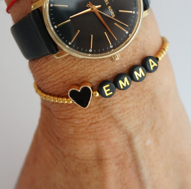 May include: A gold beaded bracelet with black letter beads spelling out the name "EMMA" and a black heart charm.