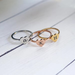 May include: Three heart-shaped rings with initial engravings. The rings are silver, gold, and rose gold. The initials are "A", "K", and "H".