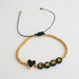 May include: A gold beaded bracelet with a black heart charm and the name "EMMA" spelled out in gold letters on black beads.