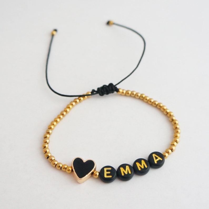 May include: A gold beaded bracelet with a black heart charm and the name "EMMA" spelled out in gold letters on black beads.