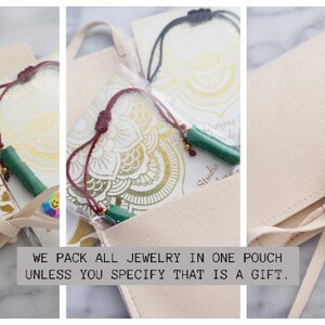May include: A close-up of a beige suede pouch with a tied ribbon closure. The pouch contains jewelry, including a green beaded necklace and a black cord necklace. The pouch is on a marble surface. The text "WE PACK ALL JEWELRY IN ONE POUCH UNLESS YOU SPECIFY THAT IS A GIFT." is printed below the pouch.
