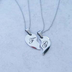 May include: Two silver necklaces with matching heart pendants. The pendants are broken in half, with one half featuring the letter "F" and the other half featuring the letter "G".
