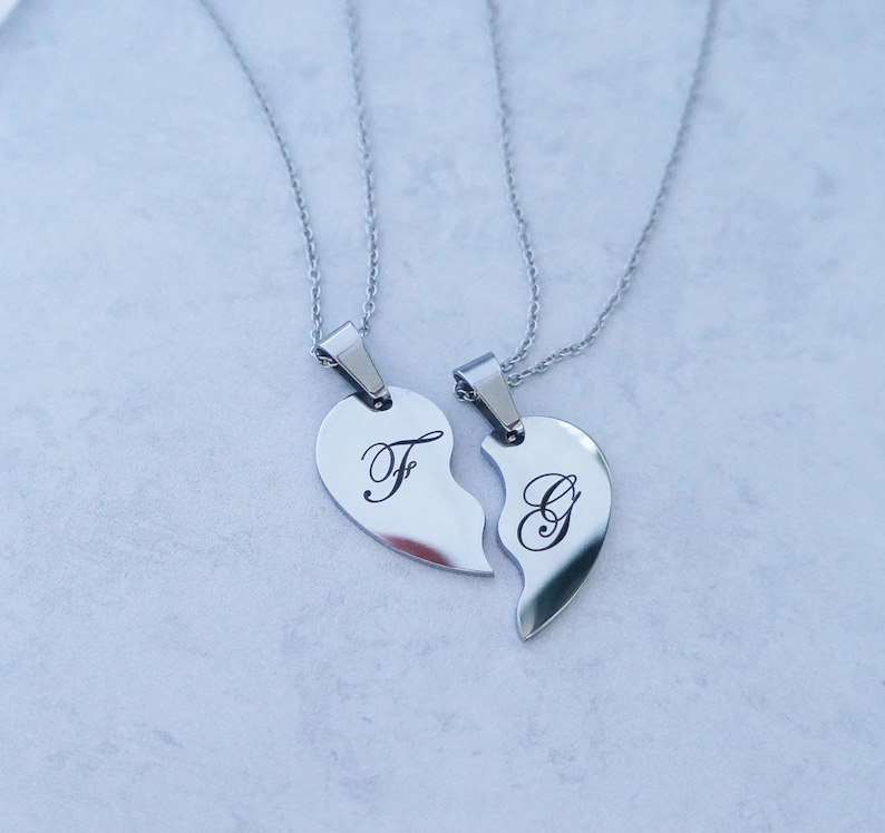 May include: Two silver necklaces with matching heart pendants. The pendants are broken in half, with one half featuring the letter "F" and the other half featuring the letter "G".