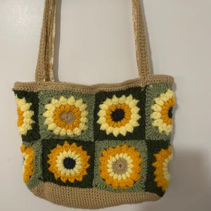 May include: A crocheted tote bag with a sunflower pattern in shades of yellow, green, and brown. The bag has two long straps.
