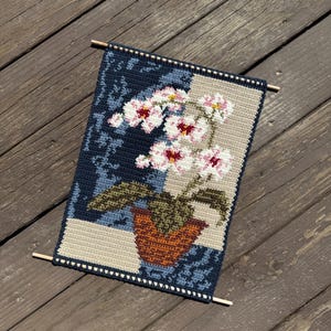 May include: Crocheted wall hanging featuring a floral design. The artwork depicts a potted orchid with white and pink flowers, set against a blue and beige background. The piece is finished with wooden dowels at the top and bottom.