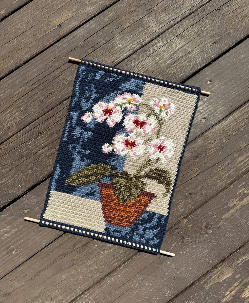 May include: Crocheted wall hanging featuring a floral design. The artwork depicts a potted orchid with white and pink flowers, set against a blue and beige background. The piece is finished with wooden dowels at the top and bottom.
