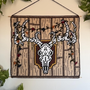 May include: A handmade wall hanging featuring a deer skull design. The skull is white and black, set against a tan background with a brown border. The artwork includes floral accents and is suspended by a wooden dowel and brown string.