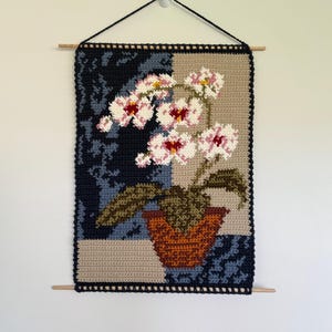 May include: A crocheted wall hanging featuring a floral design. The artwork showcases a potted orchid with white and pink flowers, green leaves, and a brown pot. The background is divided into sections of navy blue, tan, and light blue, with a black border.