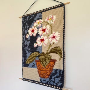 May include: A handmade crochet wall hanging featuring a floral design. The artwork showcases a potted orchid with pink and white flowers, green leaves, and a brown pot. The background is divided into sections of tan, navy blue, and patterned blue.