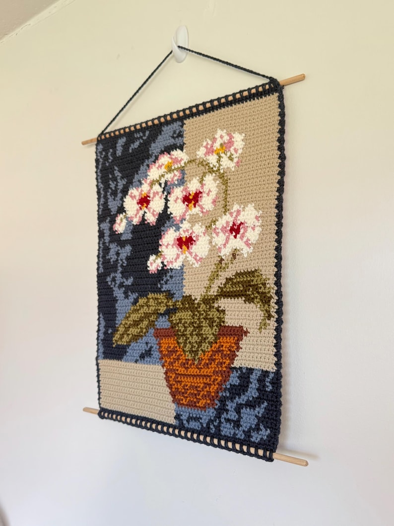 May include: A handmade crochet wall hanging featuring a floral design. The artwork showcases a potted orchid with pink and white flowers, green leaves, and a brown pot. The background is divided into sections of tan, navy blue, and patterned blue.