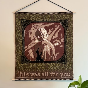 Puede incluir: Un tapiz de pared de ganchillo con una imagen central en tonos marrón, beige y negro, enmarcada por un borde oscuro. El fondo es verde moteado. El texto "this was all for you" está en la parte inferior. Colgado de una barra de madera.