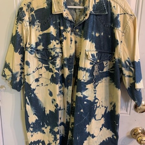 May include: A blue and yellow tie-dye denim shirt with a button-up front and short sleeves. The shirt has a faded look and a distressed design. The label on the shirt reads "Authentic Falls Creek".