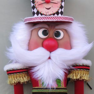 May include: A decorative Christmas nutcracker doll with a clock face on its hat. The doll has a red coat with gold trim, a white beard, and black and white checkered pants. The doll's arms and legs are red with black ball-shaped hands and feet.