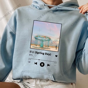 Op de afbeelding: Een lichtblauwe, getailleerde hoodie met een afbeelding van een muziekspeelerscherm. Het scherm toont het nummer "봄날 (Spring Day)" van BTS. De afbeelding is in een witte doos met een zwarte achtergrond.