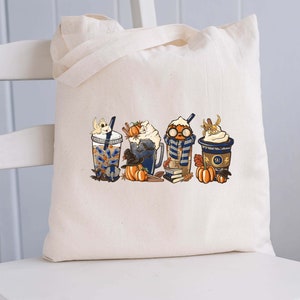 Op de afbeelding: Een witte canvas tote bag met een kleurrijke illustratie van vijf herfst-thema dranken. De dranken zijn bedekt met slagroom, karamel en pompoenspecerijen. De tas heeft een ontwerp met een pompoen, een raaf en een boek met het nummer "9 3/4" erop.